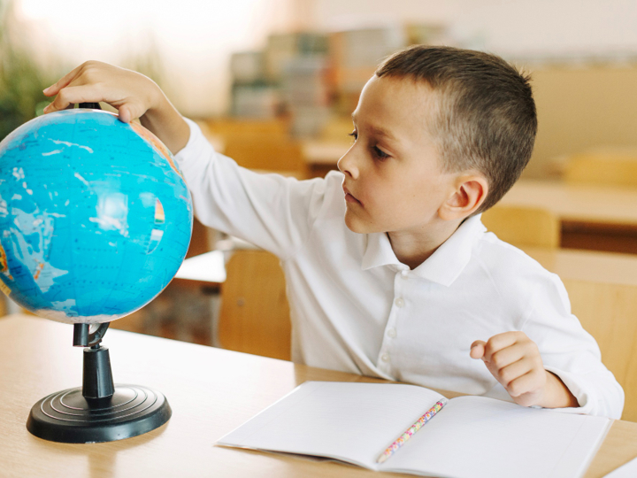 School boy in classroom sitting behind a desk with a notebook in front of him. He is turning a globe that is sitting on his desk