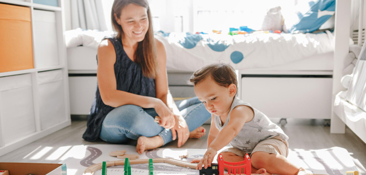 Boy toddler sitting on bedroom floor playing with train track. His mum is sitting next to him cross-legged on the floor watching him and smiling.