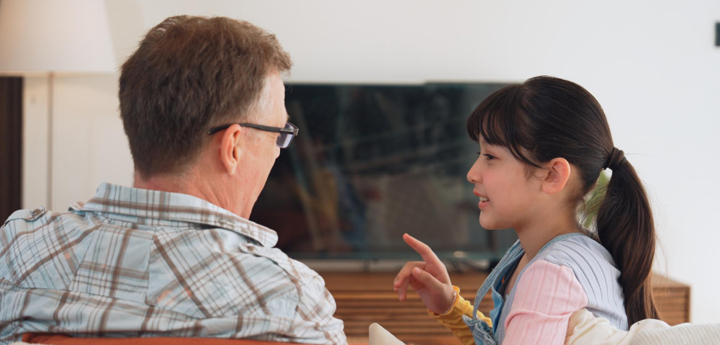 A young girl sitting on the sofa with an adult man, the girl is talking to him with one finger lifted as she speaks