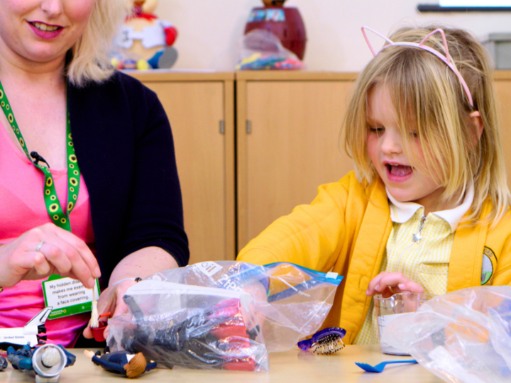 Girl in yellow cardigan and pink wire cat ears. Girl is playing with bags of plastic toys on a table. Sitting next to her is an adult helping.