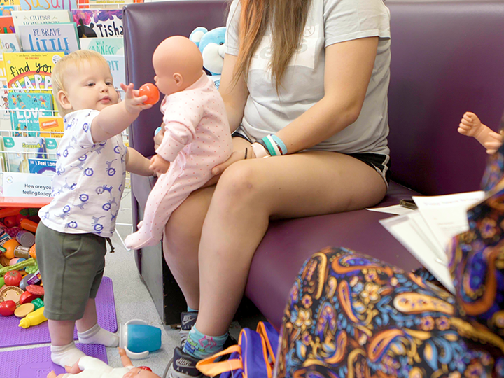 Adult sitting on a sofa with a toy doll on their lap. Toddler on the floor is standing and feeding the toy baby a toy tomato.