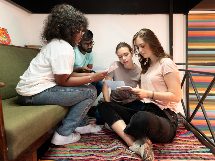 Two teenagers sitting on a sofa, whilst another 2 teenagers are sitting on the floor in front of them. All holding and looking at notes on pieces of paper