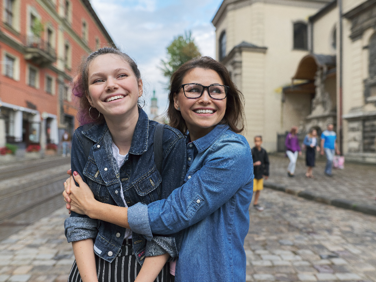A girl and a woman smiling whilst standing in the middle of a footpath. The woman is hugging the girl.