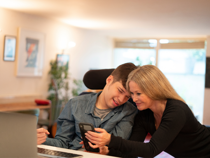 Disabled Young Adult Looking At Phone With Adult