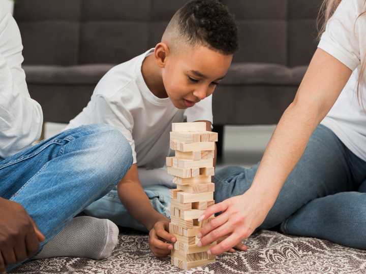 Young boy sitting on the floor playing Jenga next to 2 adults