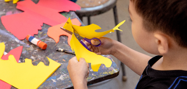 A boy cutting shapes into a yellow piece of paper with scissors.