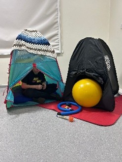 Two small tents in a classroom corner, with a mat underneath them, a sensory ball and other sensory toys. A young boy is sitting in one of the tents