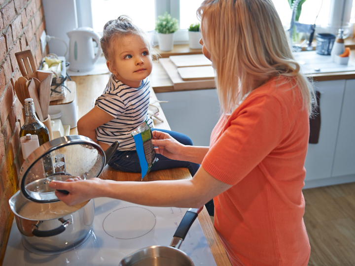 Young girl sitting on kitchen worktop looking at adult woman who is cooking on stove