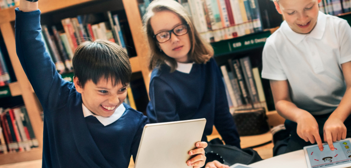 Three children in school uniform sitting in a class on ipads. One boy is raising his fit into the air with a happy and excited grin on his face