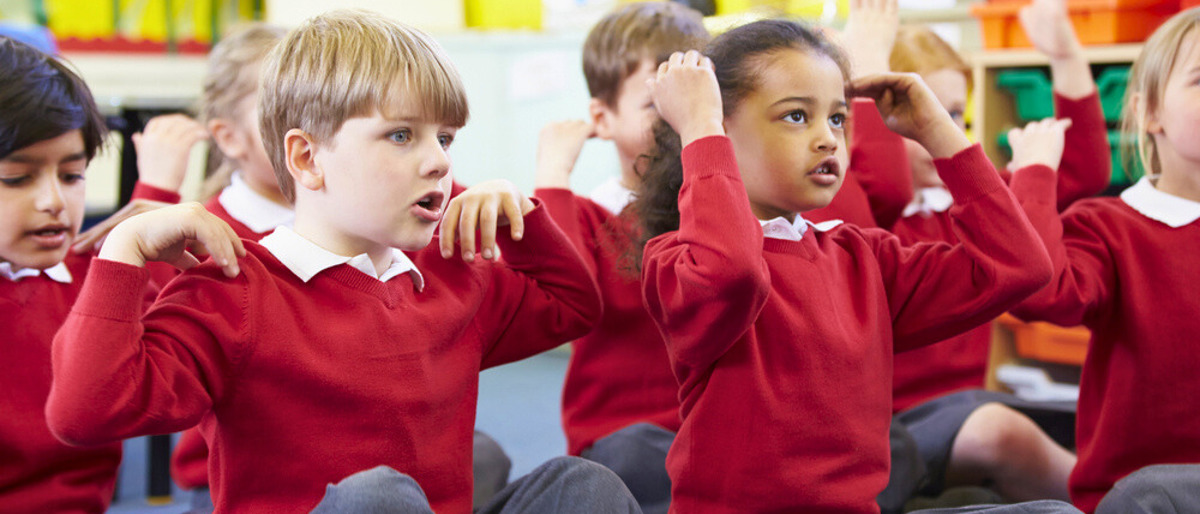 Children in classroom wearing school uniforms. The children are sitting cross-legged and singing songs with actions, touching their shoulders