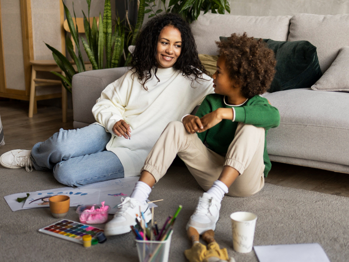 Young boy sitting on the floor leaning against a sofa, next to his mum. In front of them is colouring pens, paint and paper